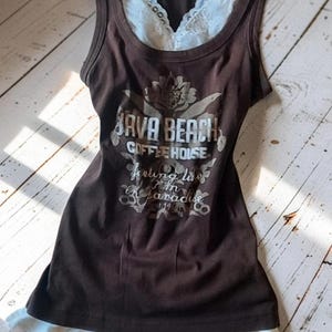 Può includere: Una canotta marrone senza maniche con bordo in pizzo bianco sullo scollo e sull'orlo. La parte anteriore presenta una grafica con il testo Java Beach Coffee House e la frase feeling like I'm in paradise, centrata sopra un motivo floreale.