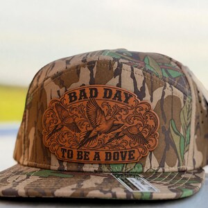 May include: A camouflage baseball cap featuring a brown leather patch on the front. The patch displays an intricate floral border, three flying doves, and the text "BAD DAY TO BE A DOVE" in a bold, dark font. The cap has a flat brim and a structured crown.
