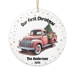 Peut inclure: Une décoration ronde en céramique blanche illustrant un pick-up vintage rose transportant un sapin de Noël et des cadeaux. Le texte indique Our First Christmas, The Andersons et 2026. Le fond est orné de petits motifs d'étoiles dorées.