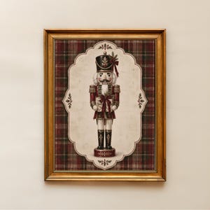 Op de afbeelding: Een ingelijst wandkunstwerk met een notenkraker-illustratie gecentreerd op een crèmekleurige geschulpte achtergrond met een donkerrode en groene ruitjesrand. De notenkraker draagt een zwarte hoed, een rood uniform en houdt een klein cadeau vast.
