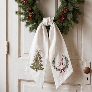 Peut inclure: Une décoration suspendue en lin crème avec deux longs pans. Un pan présente un sapin de Noël brodé en vert avec un ruban rose, tandis que l'autre affiche une lettre A rouge dans une couronne verte avec un nœud rouge.