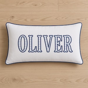 May include: A rectangular white throw pillow featuring the name OLIVER in large, bold, blue-outlined letters. The interior of each letter is filled with a light blue and white gingham check pattern. The pillow has a dark blue piped edge border.