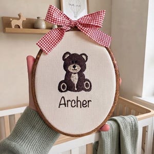 Può includere: Un telaio da ricamo ovale in legno contenente tessuto color crema con un'illustrazione di un orso ricamata in marrone scuro e il nome Archer ricamato in filo marrone scuro. Un fiocco a quadretti rossi e bianchi è fissato in cima al telaio.
