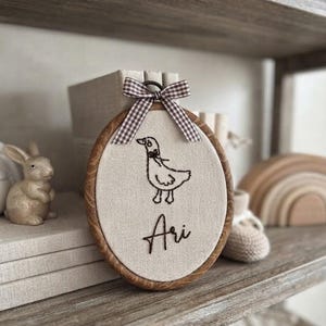 Pode incluir: Um bastidor de bordado oval com fundo de tecido creme apresentando o contorno bordado em castanho escuro de um pato com um pequeno laço. Abaixo do pato, o nome Ari está bordado em letra cursiva castanho escuro. Um laço xadrez castanho e branco está fixado.