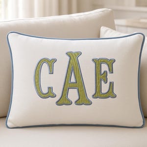 May include: A rectangular off-white throw pillow featuring the embroidered letters CAE in a textured lime green font with a light blue outline. The pillow is finished with a matching light blue piped edge along the entire perimeter of the cushion.