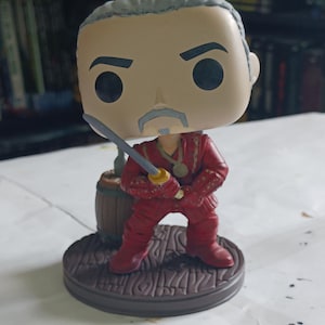 May include: A vinyl collectible figure featuring a character with gray hair and a goatee, wearing a red outfit with a gold medallion. The figure holds a gray sword and stands on a circular brown base next to a small brown barrel.