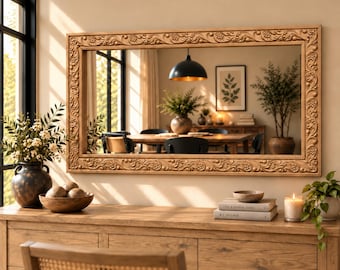 May include: A large rectangular wall mirror featuring a wide, light-brown wooden frame with intricate carved floral and vine relief patterns. The mirror reflects a dining room interior with a wooden table, chairs, and a black pendant light.