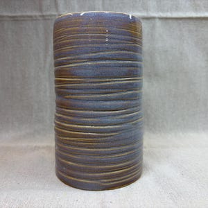 May include: A cylindrical ceramic vase featuring a textured surface with horizontal grooves. The exterior displays a blend of deep blue and earthy brown tones, with the dark blue glaze settling into the recessed lines of the pottery vessel.