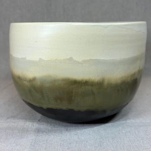 May include: A ceramic bowl featuring a smooth, rounded silhouette with a gradient glaze. The top section is a creamy off-white, transitioning into a muted olive green in the middle, and finishing with a solid dark charcoal base at the bottom.