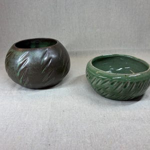 May include: Two ceramic bowls with textured surfaces. The bowl on the left is dark brown with green highlights and a rounded shape. The bowl on the right is light green with a wider, shallower opening. Both feature diagonal carved indentations.