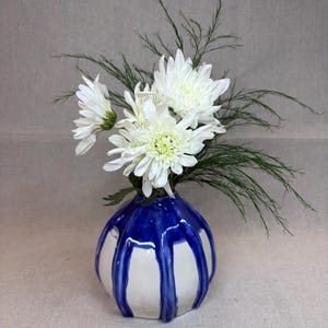 May include: A round ceramic vase with a white base and vertical blue drip glaze patterns. The vase contains a bouquet of white chrysanthemum flowers and thin, feathery green foliage, creating a contrast between the floral arrangement and the vessel.