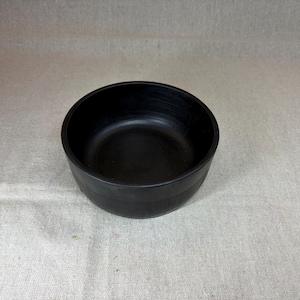 May include: A circular bowl with a matte black finish and a smooth, uniform surface. The bowl features a deep, open interior and a wide, rounded rim, presenting a simple, functional design with a consistent dark color throughout the entire object.