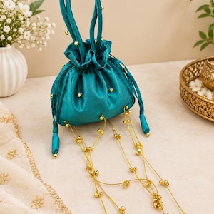 May include: A teal satin drawstring pouch bag featuring a gathered top, a single handle, and small gold beads attached to the exterior. Long, thin gold strings with clusters of small gold bells hang from the base of the bag as decorative accents.