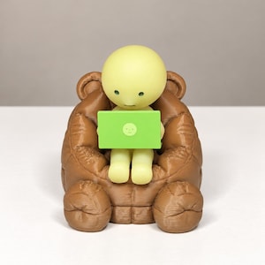 May include: A pale yellow figurine with a round head and simple facial features sits in a brown, textured bear-shaped chair. The figurine holds a bright green rectangular laptop featuring a small, circular face icon on its lid.