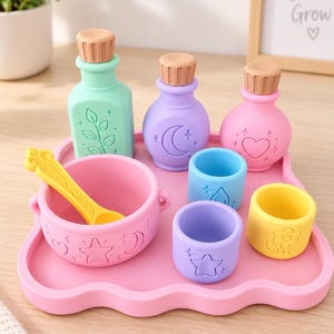 May include: A set of pastel-colored silicone play items on a pink tray, including three bottles with brown caps, a pink bowl with a yellow spoon, and three small cups in blue, purple, and yellow, all featuring embossed star, moon, and floral motifs.