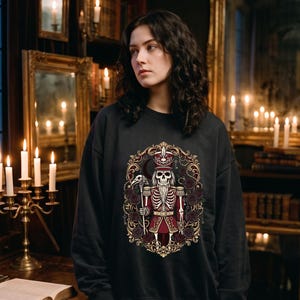 May include: A black crewneck sweatshirt featuring a graphic print of a skeletal nutcracker figure dressed in a red and gold uniform, holding a staff, surrounded by dark roses and an ornate gold frame with intricate scrollwork and key motifs.