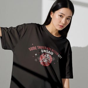 May include: A black short-sleeved t-shirt featuring a graphic of a skeleton holding a telephone. The text reads, "SOME THINGS ARE BETTER LEFT UNSAID" in an arched font, with "UNSAID" in bold, and "I'M GONNA SAY THEM ANYWAY" below the skeleton.