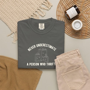 Pode incluir: Uma t-shirt cinzento-carvão com um gráfico branco de três sacos de compras e o texto "NEVER UNDERESTIMATE A PERSON WHO THRIFTS" impresso num esquema curvo. A t-shirt tem um decote redondo e uma etiqueta da marca Comfort Colors no interior.
