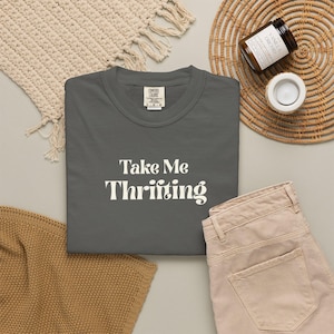 May include: A charcoal gray short-sleeve t-shirt with the white text Take Me Thrifting printed on the front. The shirt features a Comfort Colors brand label at the inner neckline and is displayed folded on a flat surface with other items.
