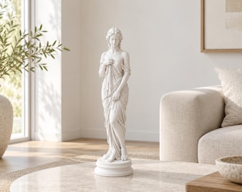 May include: A white matte finish sculpture of a standing figure draped in flowing fabric. The figure has detailed hair and is positioned on a circular pedestal base. The sculpture captures a classical aesthetic with realistic folds in the cloth.