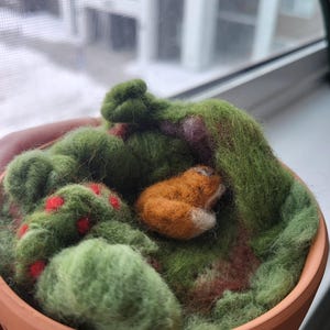 May include: A needle-felted orange fox curled up inside a terracotta bowl filled with green felted moss, brown earth-toned fibers, and small red accents. The fox has a white-tipped tail and is nestled within the textured, soft green landscape.