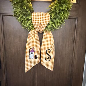 May include: A green leaf wreath adorned with a long orange and white gingham fabric ribbon. The left ribbon tail features an embroidered ghost wearing a purple hat with a black cat, while the right tail displays a large black embroidered letter S.