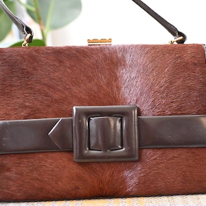 May include: A rectangular handbag featuring a brown hair-on-hide texture, accented by a wide, smooth dark brown leather horizontal strap across the centre. The strap includes a large, square, matching dark brown buckle. It has a top handle.