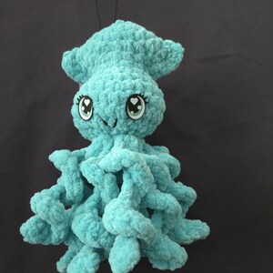 May include: A turquoise crochet plush toy shaped like a squid with a soft, fuzzy texture. It features large, round eyes with heart-shaped pupils, long eyelashes, and multiple curled tentacles extending from the base of its rounded head.