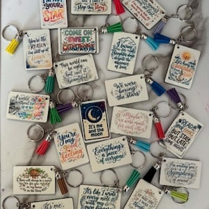 May include: A collection of rectangular keychains featuring various inspirational quotes and colorful illustrations, each attached to a metal ring and a small decorative tassel in assorted colors like yellow, red, blue, green, purple, and black.