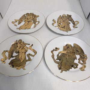 May include: A set of four white ceramic plates, each featuring a central gold-toned illustration of an angel playing a different musical instrument. Each plate is finished with a thin gold rim, showcasing detailed wings and flowing ribbon motifs.