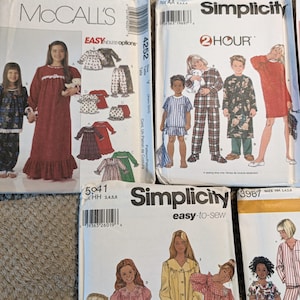 May include: A collection of four paper sewing pattern envelopes from McCall's and Simplicity brands. The envelopes feature illustrations of children and adults wearing various styles of pajamas, nightgowns, and loungewear, with printed text.