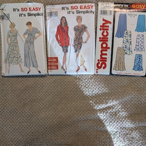 May include: Three Simplicity sewing pattern envelopes labeled It's SO EASY. The left envelope shows a floral dress and a plaid jumpsuit. The center envelope displays a red jacket and a floral dress. The right envelope features various skirt designs.