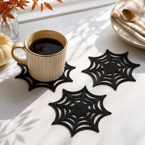 Peut inclure: Trois dessous de verre en feutrine noire en forme de toile d'araignée. L'un des dessous de verre supporte une tasse en céramique beige à côtes verticales remplie de café noir. Ils présentent des motifs découpés avec un centre et des rayons.