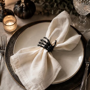 Peut inclure: Un rond de serviette en forme de main de squelette noire enserrant une serviette en tissu blanc cassé. Le rond de serviette représente une main humaine aux doigts articulés, posé sur une assiette blanche à bordure dorée sur un sous-plat.