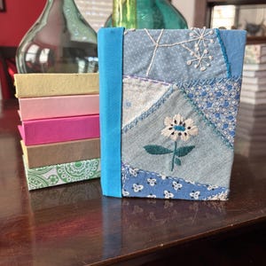 May include: A fabric-covered journal featuring a patchwork design of various blue patterned textiles, accented with white embroidered floral motifs and a solid blue spine, standing beside a stack of five small, colorful rectangular boxes.