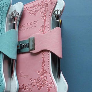 May include: Two rectangular journals, one light blue and one pink, featuring embossed floral patterns, a silver metal combination lock closure, and a silver pen with a crown-shaped topper. The text "BLUEPRINT" is printed on the front covers.