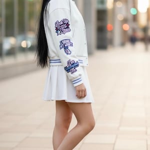 May include: A white varsity jacket featuring blue striped ribbed cuffs and collar, adorned with multiple embroidered patches on the sleeve including the text Look Up, I Shine My Light, and I am Unapologetically Me, paired with a white pleated skirt.