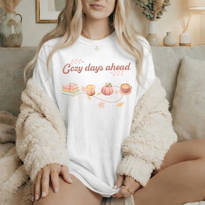 Puede incluir: Una camiseta blanca con el texto "cozy days ahead" en una fuente cursiva marrón, acompañada de una ilustración gráfica de una pila de libros, una vela encendida, una calabaza rosa y una taza de café, conectadas por una fina línea marrón.