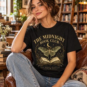 May include: A black short-sleeved t-shirt featuring a gold and green graphic of an open book, a moth, and a crescent moon. The text reads THE MIDNIGHT BOOK CLUB, ONE MORE CHAPTER, and GALLERY MOOD STUDIOS. The design includes leafy vine accents.