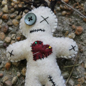 May include: A white felt voodoo doll with black stitching, a red felt heart with pins, and a black button eye. The doll is lying on a bed of small rocks.