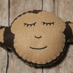 May include: A handmade monkey-shaped plush toy. The monkey has a light brown face with closed eyes and a smiling mouth, and dark brown ears and head outline. The toy is made of felt and stitched together with a contrasting thread.