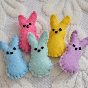 May include: A collection of five colorful felt bunny ornaments. The bunnies are in shades of pink, yellow, blue, mint green, and purple. Each bunny has black bead eyes and a stitched outline. These Easter decorations are perfect for spring.