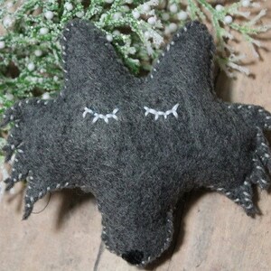 May include: A handmade gray felt wolf ornament with white stitched details. The wolf has closed eyes and a black nose. The edges are stitched with white thread. Decorative greenery is in the background.