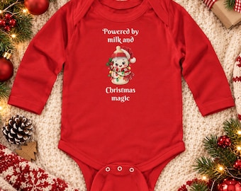 Puede incluir: Un body de bebé rojo de manga larga con un gráfico de un biberón disfrazado de muñeco de nieve con gorro de Papá Noel y bufanda. El texto Powered by milk and Christmas magic aparece en letras blancas sobre y bajo el gráfico.