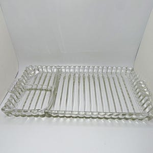 May include: A rectangular clear glass serving tray featuring a scalloped edge and a ribbed texture throughout the surface. The tray is divided into two sections, with a smaller square compartment on the left and a larger rectangular area on the right.