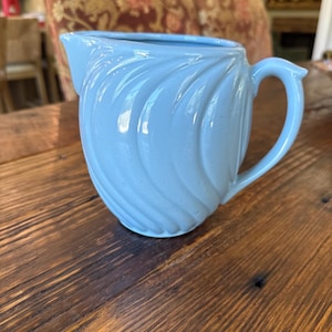 May include: A light blue ceramic creamer pitcher featuring a glossy finish and a distinct swirling, ribbed texture across its rounded body. The vessel includes a curved handle on the side and a small pouring spout at the top rim for liquid dispensing.