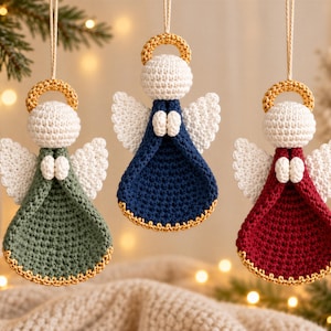 Op de afbeelding: Drie gehaakte engelhangers die aan touwtjes hangen. Elke engel heeft een wit hoofd, witte vleugels, kleine witte handjes en een gouden halo. De jurken zijn saliegroen, marineblauw en donkerrood, elk afgewerkt met een goudkleurige rand.