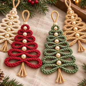 four crocheted Christmas tree ornaments in various colors, including red, green, and beige, arranged on a beige cloth. The ornaments are surrounded by pine branches and pine cones, creating a festive holiday scene.