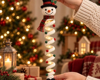 Op de afbeelding: Een gehaakte sneeuwpop-ornament met een zwarte hoge hoed, een rood-groen gestreepte sjaal en een lang, spiraalvormig wit lichaam versierd met kleine rode en groene pompons. Het spiraalvormige lichaam bevat geïntegreerde warmwitte LED-verlichting.