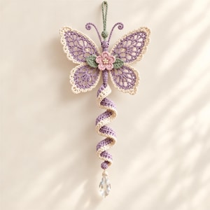 Puede incluir: Un adorno colgante de mariposa hecho a ganchillo con alas de encaje en color lavanda y crema, una flor rosa central con hojas verdes y una cola en espiral lavanda y crema que termina en un colgante de cristal transparente y facetado.